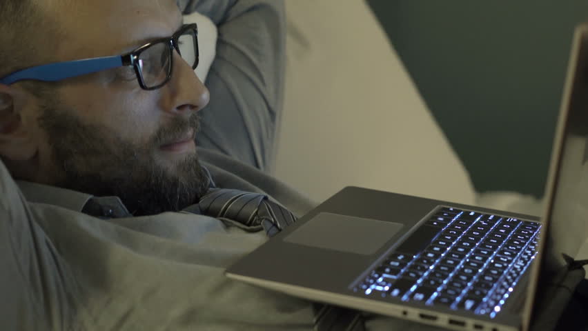 Young businessman watching movie on laptop on bed in hotel room at night