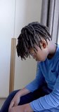 Vertical video of sad african american boy sitting next to window at home, slow motion. Childhood and domestic life, unaltered.