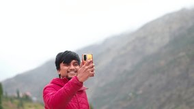 Portrait of a happy young Indian man waving his hand while holding his mobile phone during his video call in holidays at Manali in Himachal Pradesh, India. Happy man talking on video call via mobile.