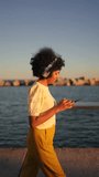 Video of the side view of an african woman using phone while walking with headphones in the promenade next to the sea
