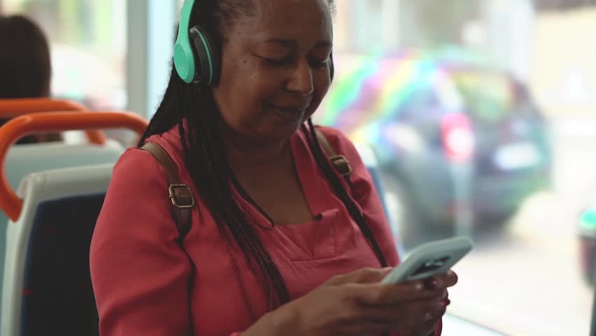 Happy senior african woman using mobile phone while listening music playlist inside tram transportation - Joyful elderly and travel concept Royalty-Free Stock Footage #3463757579
