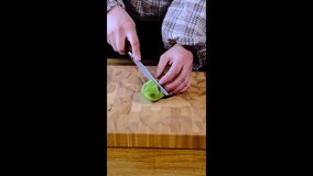 A woman in a checkered shirt and jeans cuts a peeled kiwi on pieces on a wooden board. Video recipes. Cooking
