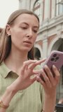 Vertical video, Beautiful woman with brown hair, dressed in an olive green sweater, uses a map application on her mobile phone while standing in the old town square