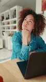Vertical video, Young woman with curly hair wearing denim shirt with laptop sitting at table, looking up, thinking at home