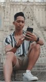 VERTICAL VIDEO: Young man tourist on steps uses mobile phone while sitting on landmarks background