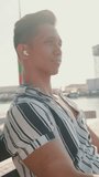 VERTICAL VIDEO: Young man tourist sits on bench on the pier, listens to music. Backlight