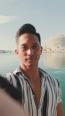 VERTICAL VIDEO: Close-up of young man standing on dock taking selfie with cell phone