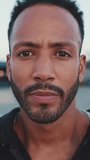 VERTICAL VIDEO: Young bearded male athlete standing on the embankment, looking at the sea. Guy turns his head and looks at the camera. Camera moving forwards