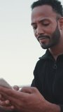 VERTICAL VIDEO: Close up, young smiling bearded male athlete dressed in hoodie uses cellphone after training while sitting on the embankment