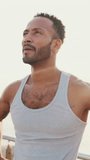 VERTICAL VIDEO: Young bearded male fit athlete standing on the embankment against the sea background, looking at the camera
