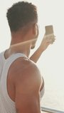 VERTICAL VIDEO: Young bearded male fit athlete taking photo on mobile phone relaxing after workout on the embankment. Back view. Backlight