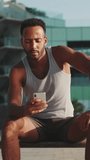 VERTICAL VIDEO: Young bearded male athlete is resting after training, sitting on bench, using smartphone on modern building background
