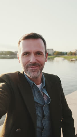 VERTICAL VIDEO: Close up, man makes video call on mobile phone on the embankment of the river. Backlight