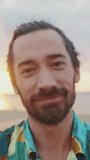 VERTICAL VIDEO: Close-up of young smiling man looking at the camera on the beach at sunrise