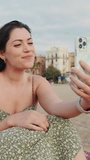 VERTICAL VIDEO: Happy young woman making video call while sitting on the beach