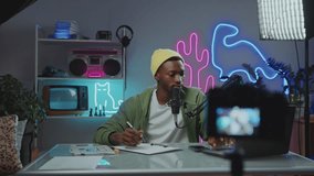 Young African American blogger using laptop and taking notes when preparing content plant at workplace in modern video studio with neon decor, light setup and camera