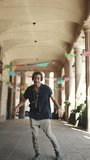 VERTICAL VIDEO: Young italian guy with ponytail and stubble in headphones goes to the city landscape background. Stylish guy listens to music in headphones and dances