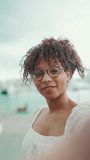 VERTICAL VIDEO: Closeup of a young woman in glasses stands in the seaport and shots selfie on a smartphone and blows a kiss. Positive woman using mobile phone outdoors in urban background.
