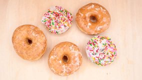 4K HD video zooming in on traditional round glazed donuts and vanilla frosted donuts with rainbow sprinkles, laying on a light wood table.