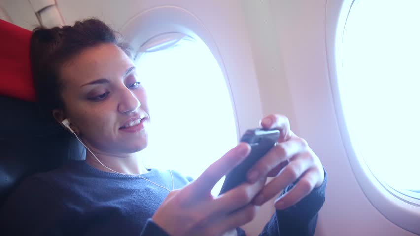 Woman on a plane listening to music with her smartphone - Travel and technology concepts, young woman wearing noice cancelling earphones