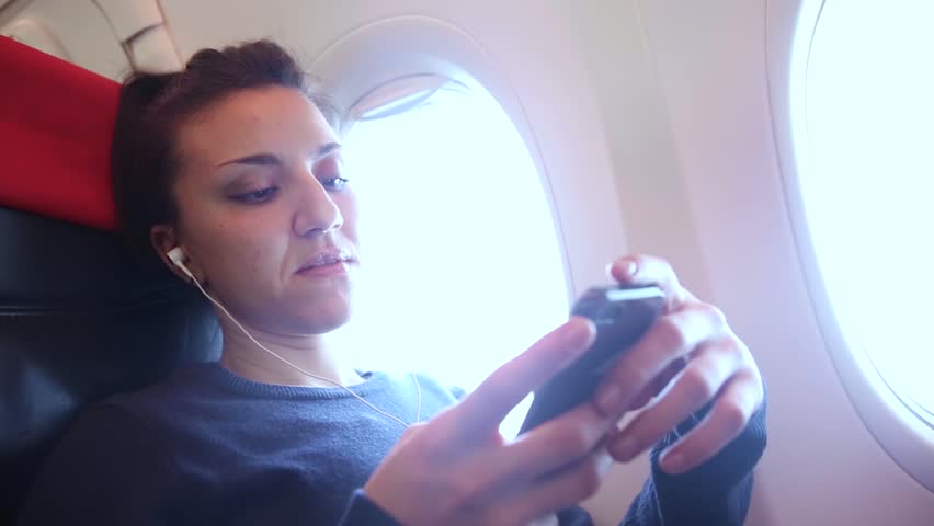Woman on a plane listening to music with her smartphone - Travel and technology concepts, young woman wearing noice cancelling earphones