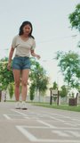 Vertical video: Joyful beautiful brunette girl dressed in casual clothes playing hopscotch game outdoors, having fun