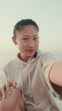 VERTICAL VIDEO, Smiling girl wears sportswear sits on the promenade, takes selfie at morning time on the sea background