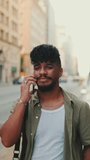 VERTICAL VIDEO: Young happy man with beard dressed in an olive-colored shirt is talking on cellphone standing on the old city background