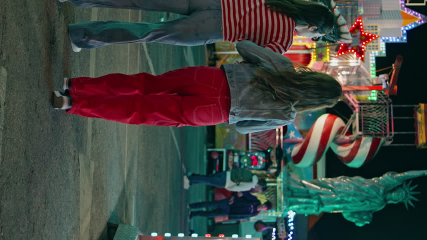 Teenage girls walking amusement park rear view. Happy best friends having fun enjoy summer holiday with illuminated carousel vertically. Unrecognized excited sisters going funfair. Weekend leisure