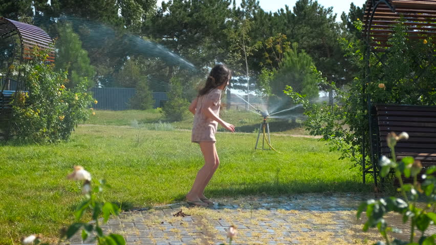 Moment of child fun with water sprinkles. A view of little girl happy moments in the automatic water sprinkle in the sunny park during summer time.