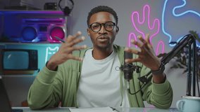 Young Black man sitting at desk with laptop and microphone and speaking on camera when filming content for video blog in studio with trendy neon light. Zoom-out shot