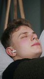 Vertical video. Cozy young blonde man with wireless in-ear headphones enjoys listening to music his eyes closed while lying on the sofa at home