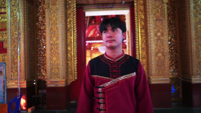 A Thai man, dressed in respectful clothing walks with a calm demeanor inside a vibrant Buddhist temple. The camera follows him as he moves towards a golden Buddha image.