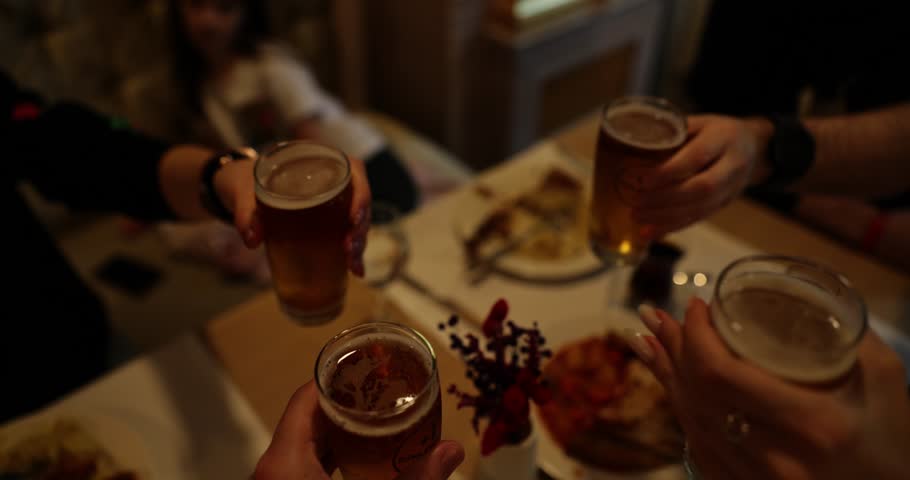 Close friends clink transparent glasses with beer in joyful semi-dark atmosphere. Cheerful pals gather to celebrate special event slow motion