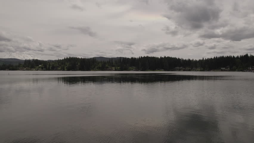 Ungraded aerial footage glides over a placid freshwater lake in Washington State, capturing its tranquility beneath a blanket of clouds.