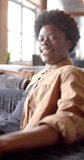 Vertical video of portrait of happy african american man sitting on sofa at home, slow motion. Lifestyle and domestic life, unaltered.