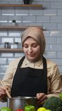 Vertical video. Beautiful young woman with headscarf cooks soup at home in modern kitchen and checks its taste