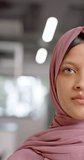Vertical video of half portrait of biracial casual businesswoman in hijab in office, slow motion. Casual office, business and work, unaltered.