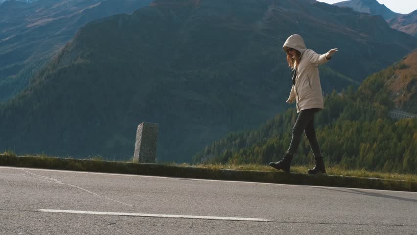 Beautiful woman traveler walks balancing on the curb and enjoying the incredible views of the mountains. Majestic Grossglockner Mountain Road in Austria, snow covered sharp peaks on background.