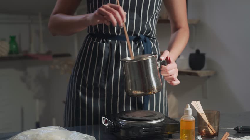 A woman in a striped apron makes wax candles. She stirs the melted wax in a pan.The creative process of making wax scented candles.