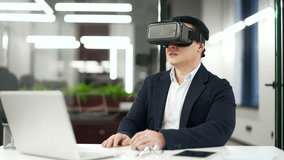Asian businessman in a formal suit works using VR glasses in virtual reality simulator sitting in business office. Manager uses gestures to control computer programs and flips through virtual pages