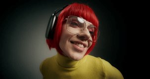 A woman with glasses, an orange wig and headphones, dancing and singing standing on a gray background in the studio, close-up of her face