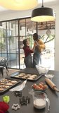Vertical video of happy diverse couple baking christmas cookies dancing in kitchen, slow motion. Cooking, fun, togetherness, christmas, tradition, celebration, domestic life and lifestyle, unaltered.