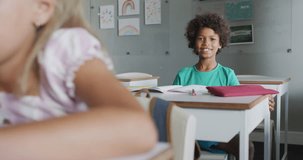 Video of happy biracial boy sitting at desk in classsroom. primary school education and learning concept.