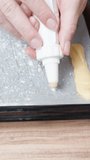 The woman is preparing eclairs. She is piping choux pastry onto a baking sheet, in a close-up shot. Vertical video.