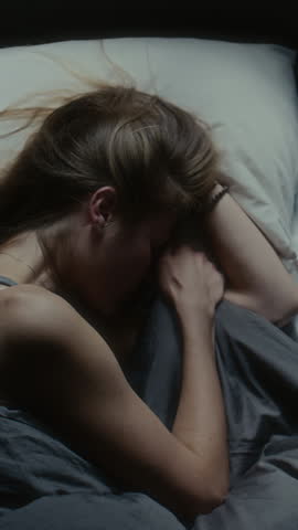 Vertical top down shot of girl lying in bed at night, gripping her hair nervously and crying