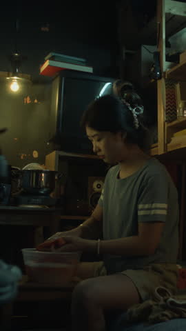 Full vertical shot of impoverished Asian or Korean girl in t-shirt and shorts sitting in dimly lit shabby tiny bedsit apartment in evening, washing clothing by hand in small plastic tub of soapy water
