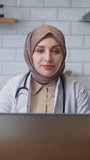 Vertical video. Female doctor in headscarf wears white coat talking to camera while sitting in kitchen