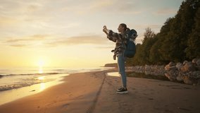 Woman tourist takes photo of sea sunset. Female hiker camper with touristic backpack recording video of nature on smartphone enjoying vacations. Travel tourism vacation camping wanderlust concept.