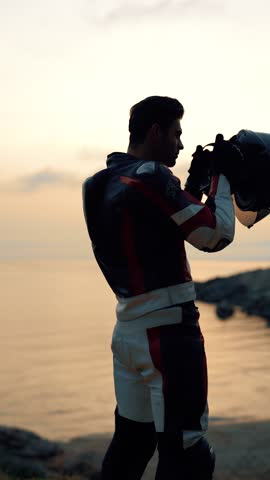 A biker puts on a helmet against the backdrop of the sea and the setting sun. Vertical video.